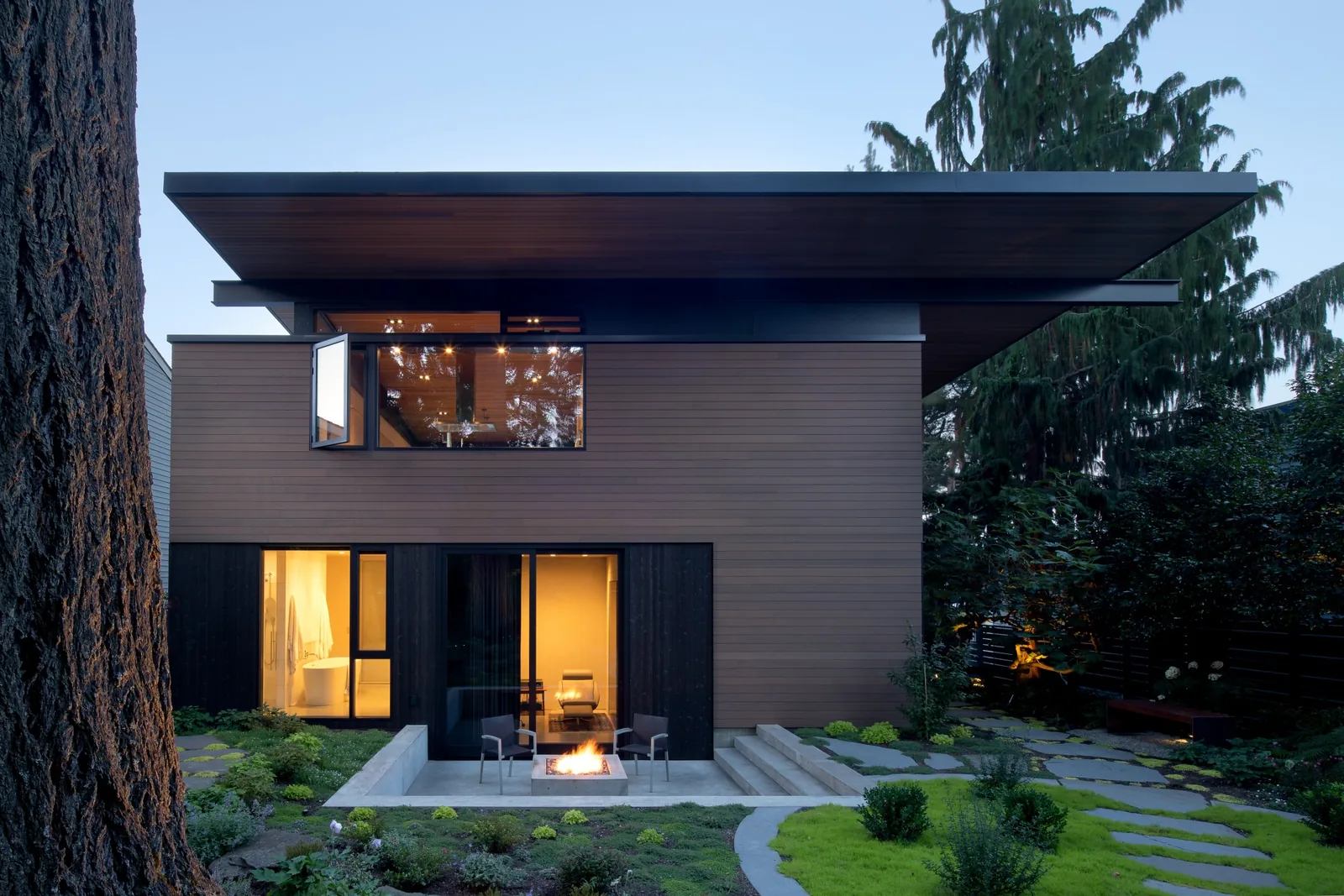 Rear facade of a modern home featuring wood siding, large windows, and a recessed courtyard with a fire pit.