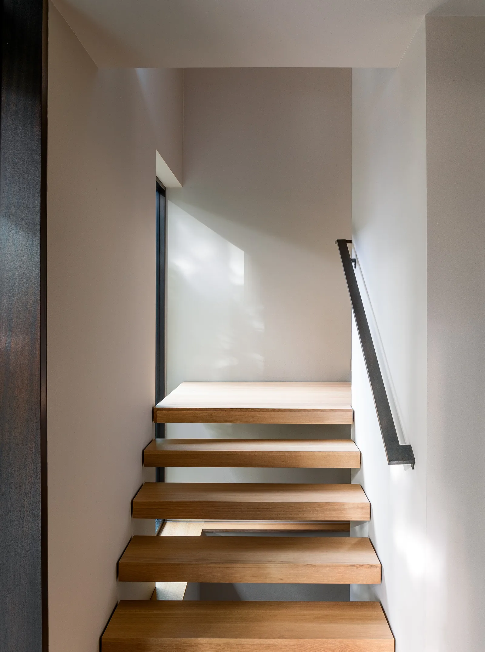 Floating wood staircase with metal handrail set against white walls and natural light from a side window.