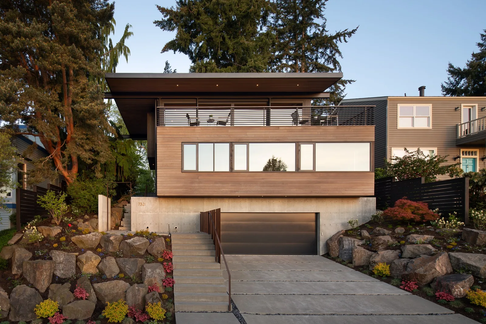 Modern house with wood siding, concrete foundation, flat roof, and large windows with balcony above garage.