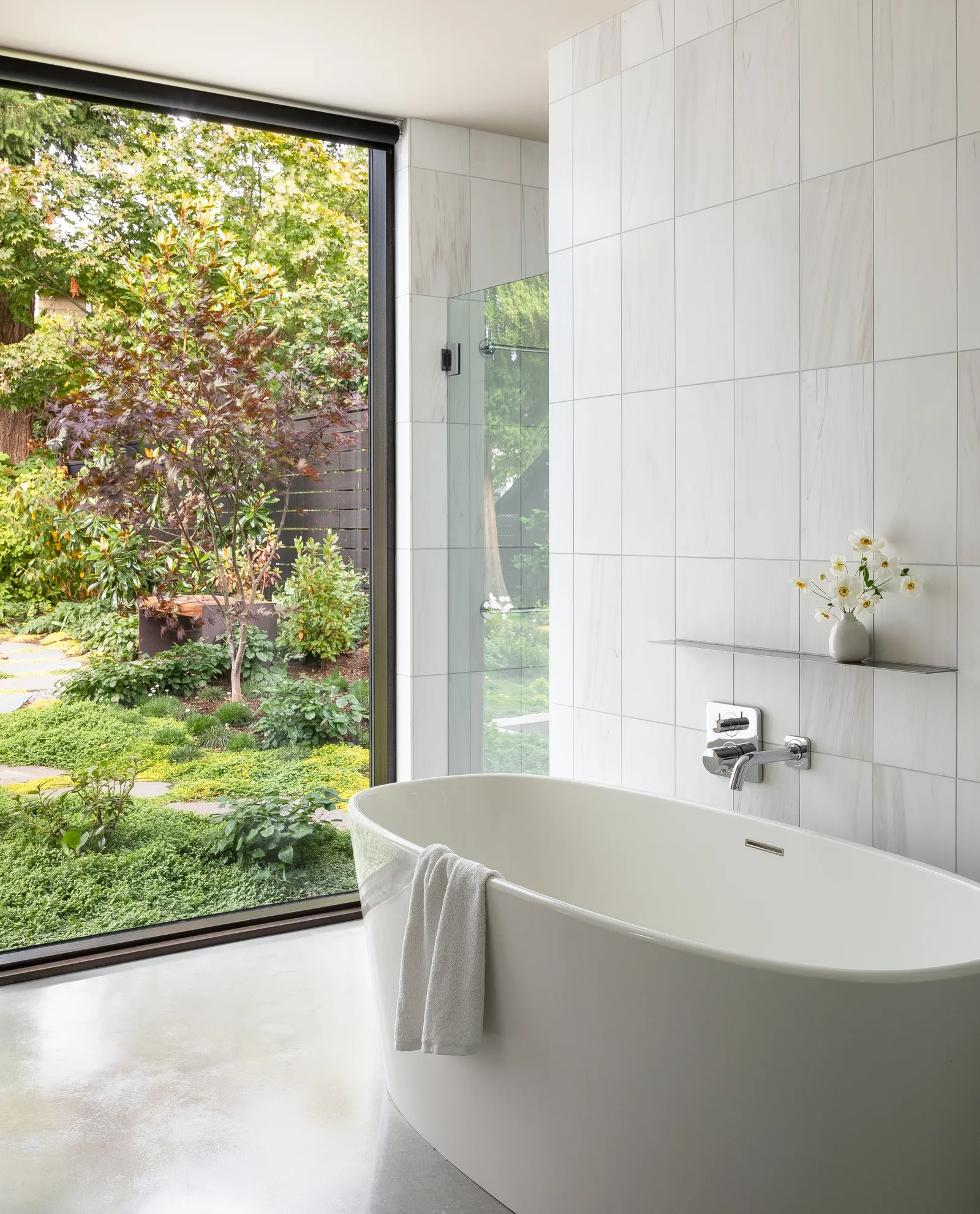 A freestanding bathtub sits adjacent to a large floor-to-ceiling window with a view of a landscaped garden, in a bathroom with tiled walls.