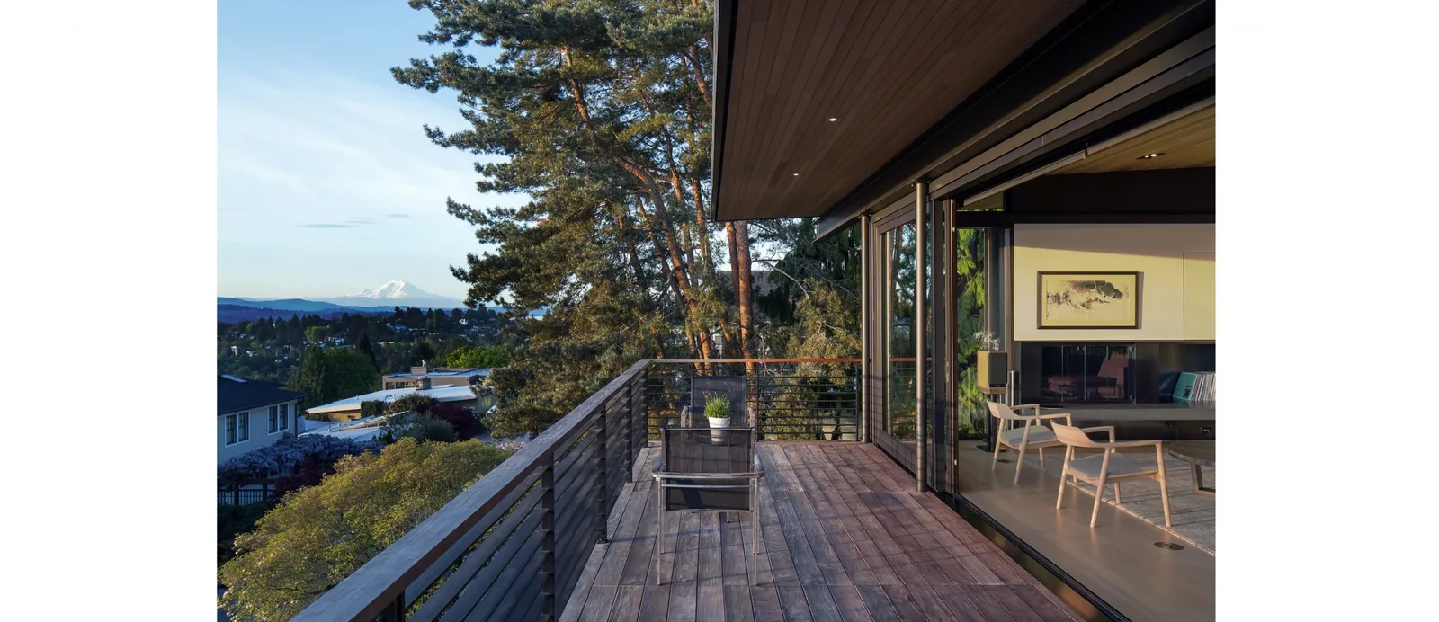 Wood deck adjacent to a living room with folding glass doors and railing overlooking a forested sloped site and distant mountains.