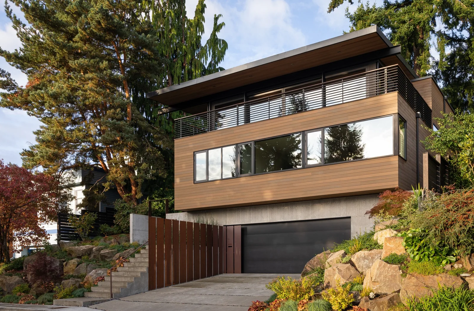 Exterior front view of a modern house featuring wood siding, flat roof, large horizontal windows, concrete base, and stone landscaping.