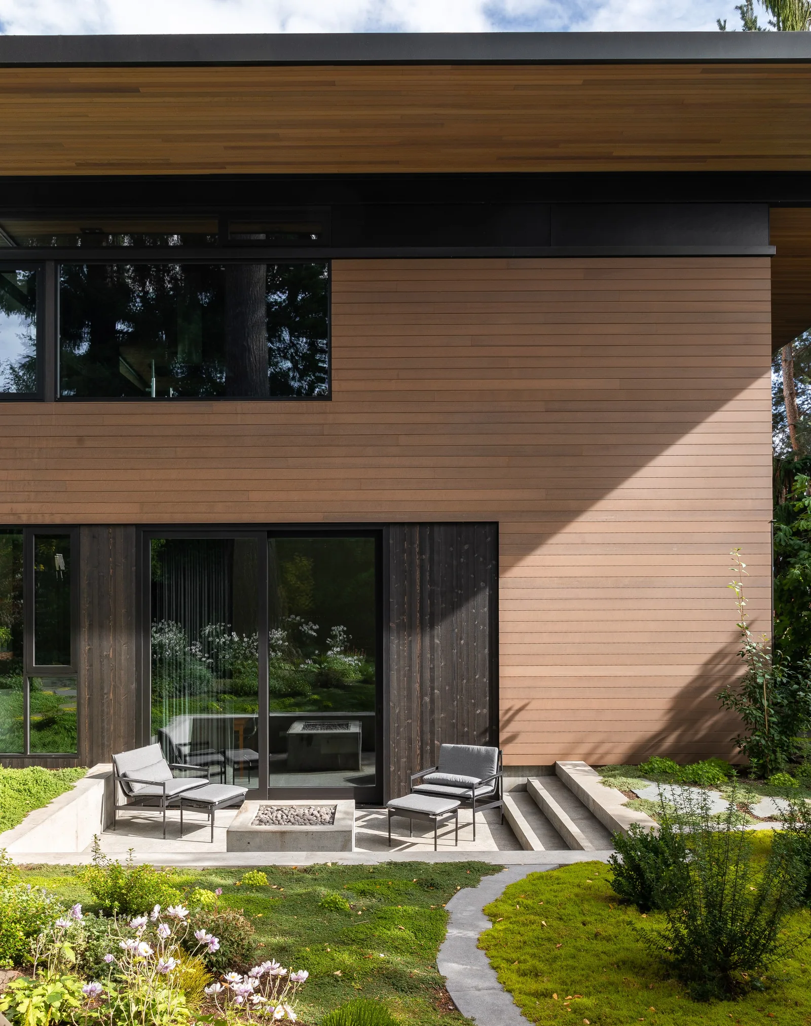 Rear exterior view of the house with wood siding, recessed concrete patio, and large sliding glass doors.