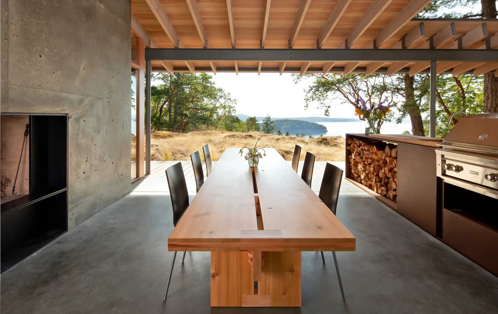 Outdoor dining space with wooden pergola, concrete flooring, built-in fireplace, and integrated grill, overlooking a natural landscape.
