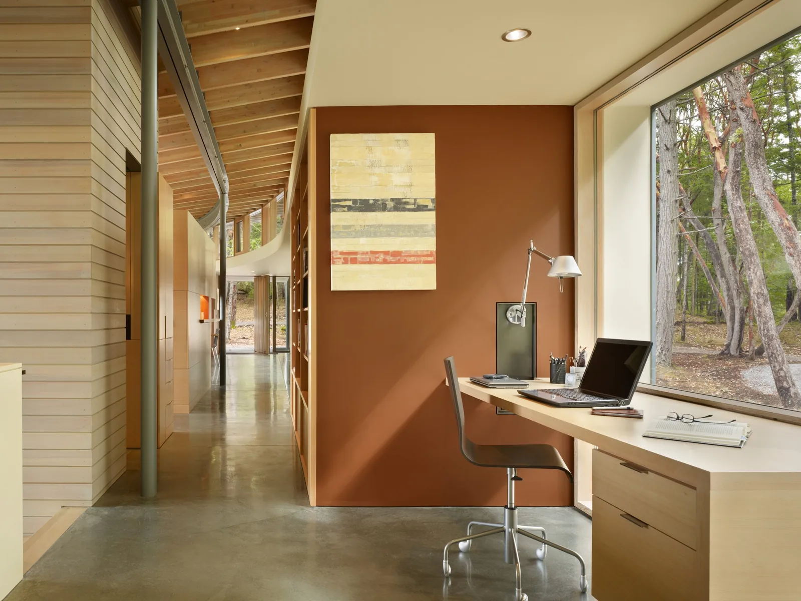 A home office nook with a built-in wooden desk and large picture window adjacent to a hallway with exposed wood ceiling and polished concrete floor.