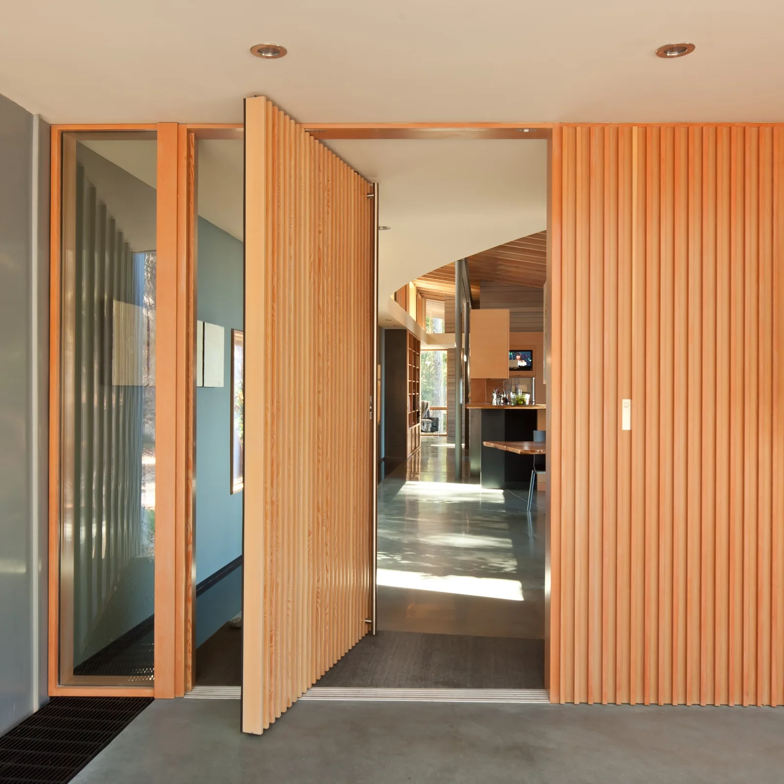 View through an open vertical slat pivot door showing interior hallway and kitchen area with polished concrete floor and wood detailing.