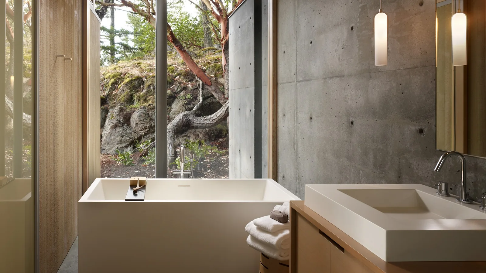 Bathroom with concrete wall, freestanding bathtub, rectangular sink, and large windows overlooking rocky terrain.