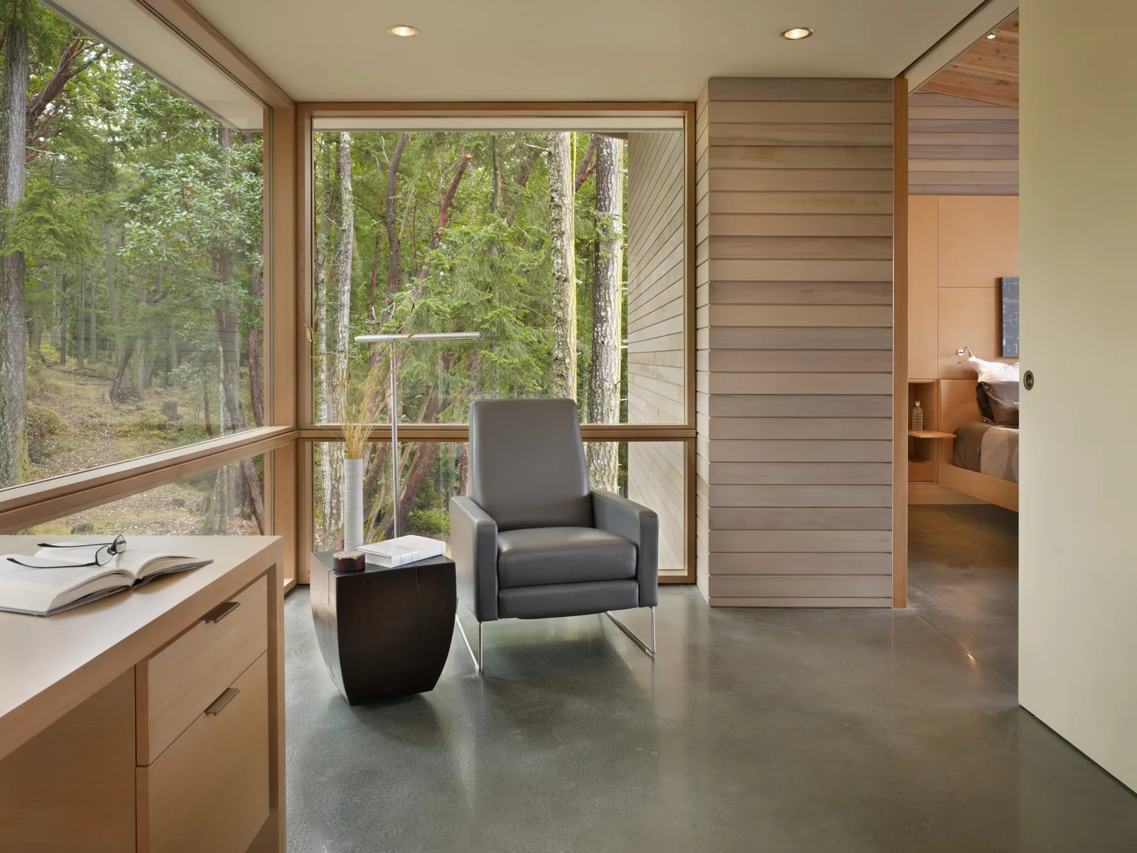 Corner of a room with large windows overlooking a forest, featuring a modern armchair and side table, adjacent to a bedroom.