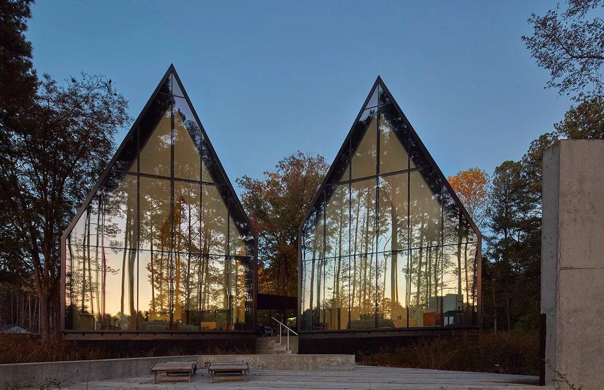 Steeplechase House featuring two symmetrical steep-pitched glass volumes with black framing reflecting the wooded surroundings.