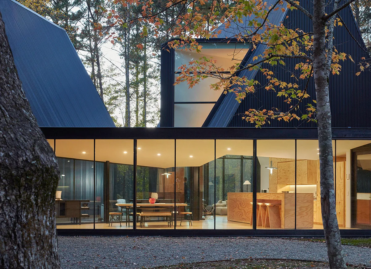 The Steeplechase House features a black angled metal roof with full-height glass walls and wood-paneled interiors.