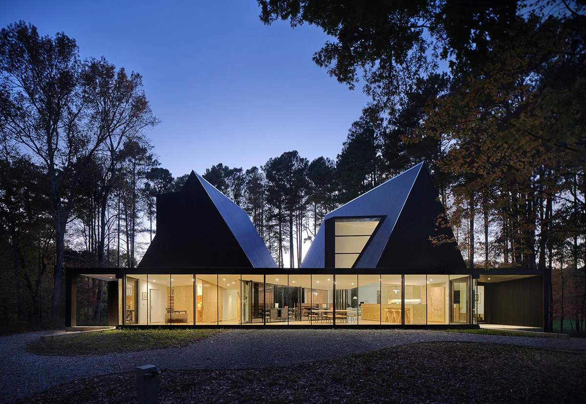 Exterior front view of the Steeplechase House with illuminated interior at dusk, featuring two steep gable roofs and floor-to-ceiling glass walls.