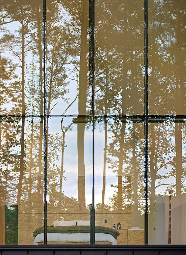 Bedroom with a bed and wood-paneled walls viewed through a large floor-to-ceiling glass window reflecting surrounding trees.
