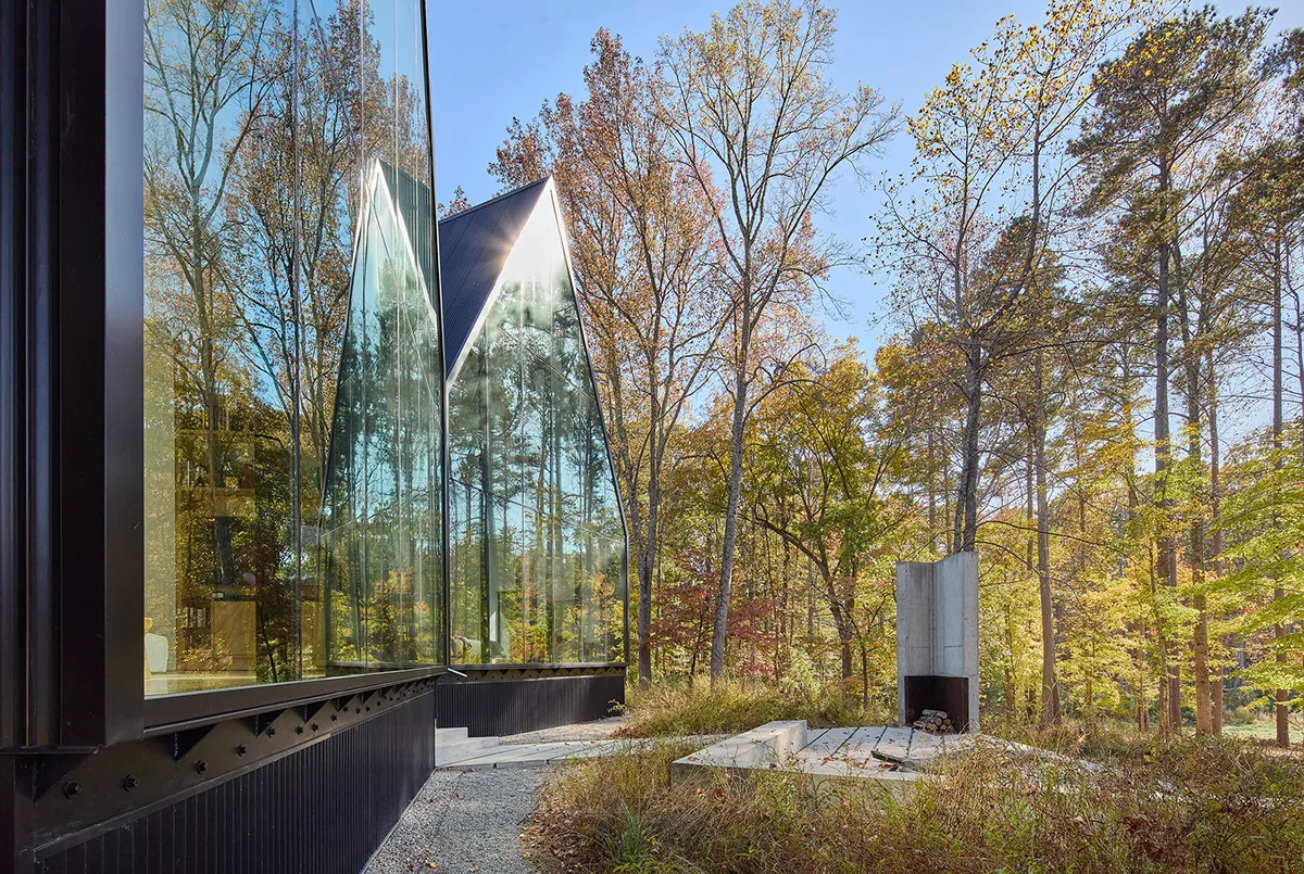 Side exterior of the Steeplechase House with reflective glass cladding and a concrete outdoor fireplace in a natural wooded setting.