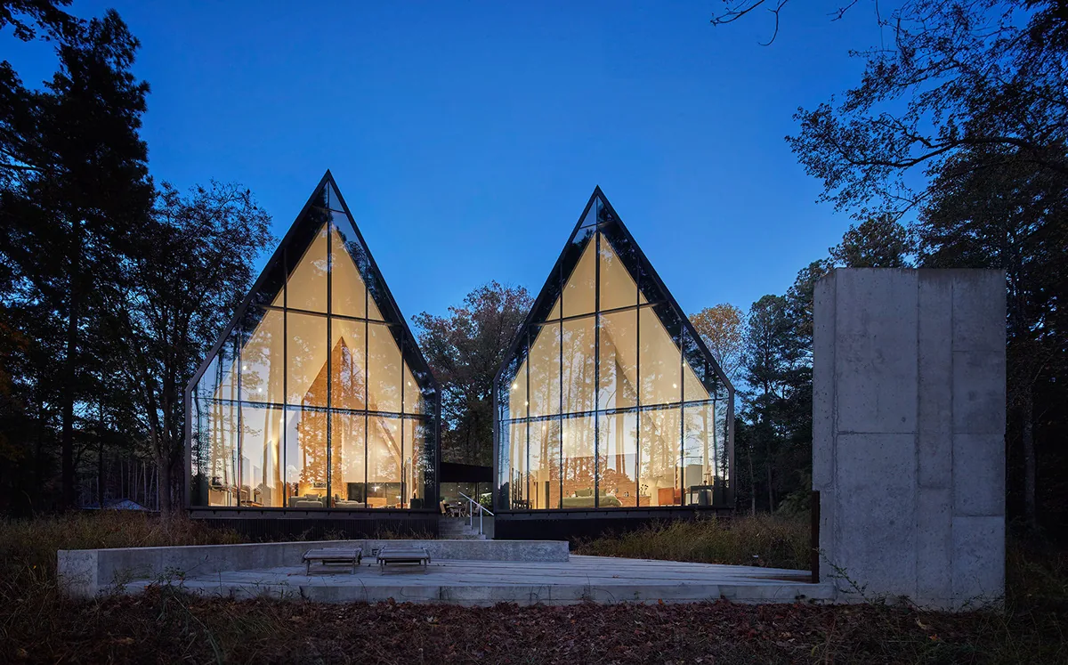 Two modern A-frame glass houses with steep roofs and concrete platform in front, shown at dusk in wooded area.