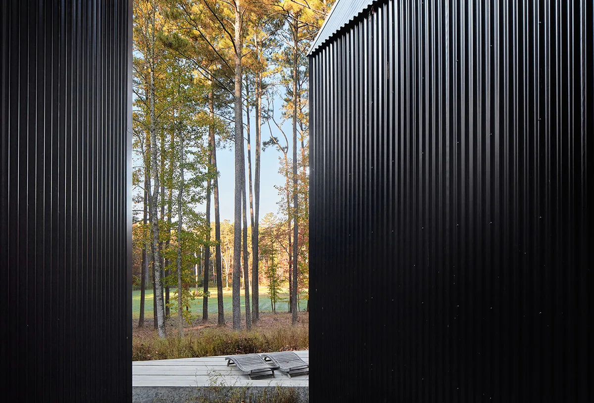 Narrow exterior space between black corrugated metal walls with view to trees and patio seating.