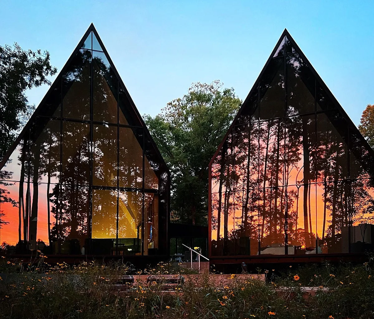 Twin A-frame structures with floor-to-ceiling glass walls reflect a sunset and surrounding forest.