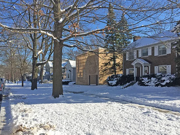 Snow covered front lawns and leafless trees line a residential street with a wooden cube-shaped building beside a brick house.