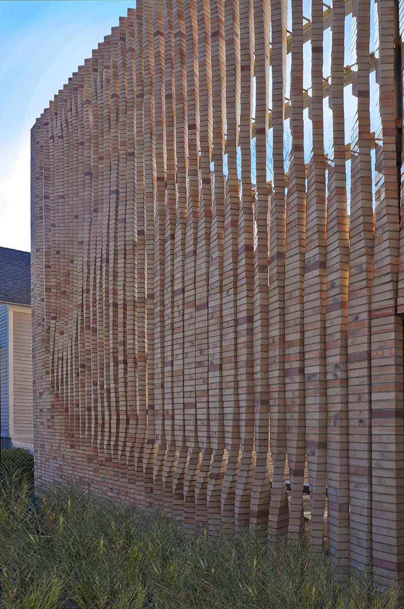 Brick wall with vertical slat design creating a textured facade and light gaps, surrounded by dense green grasses.