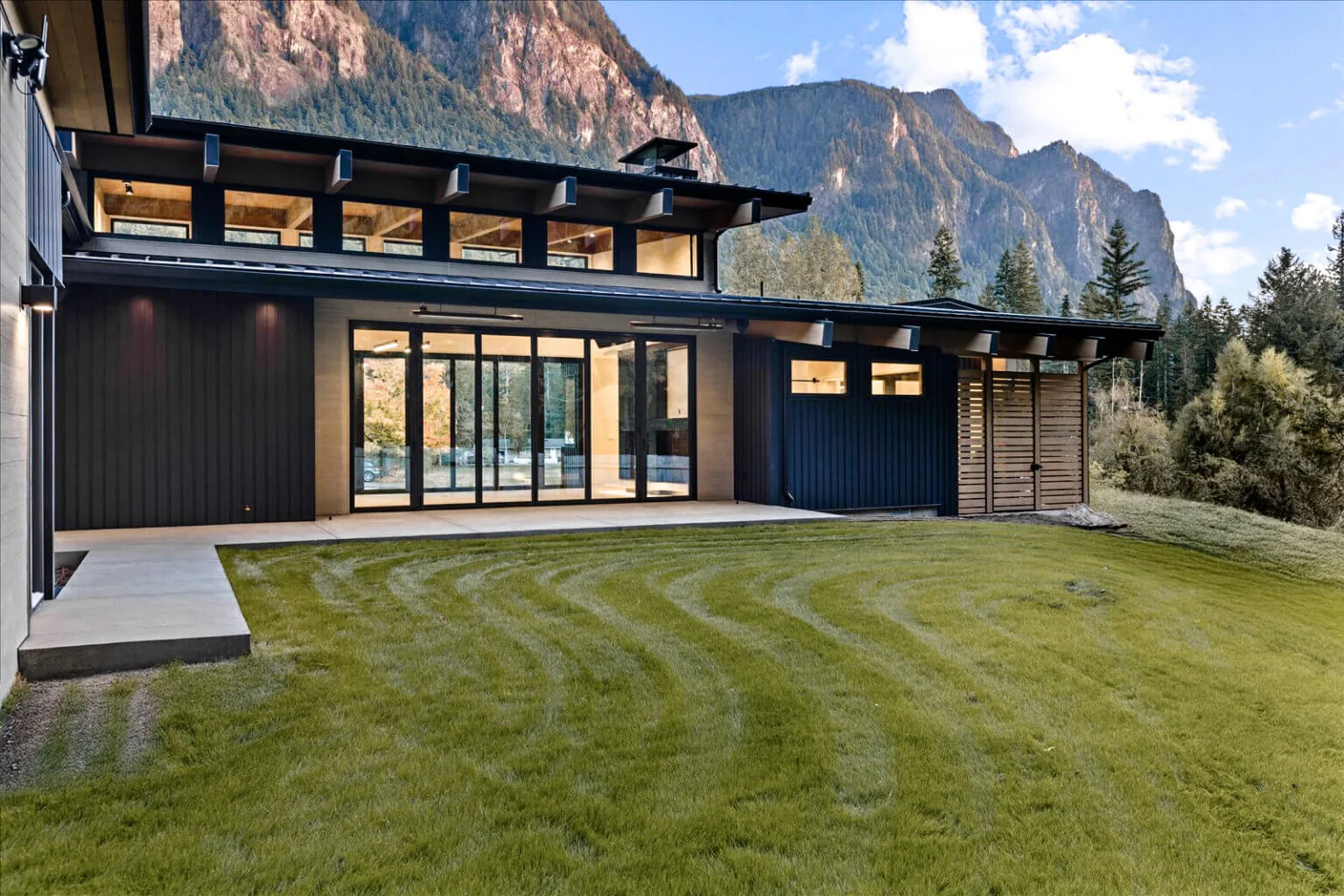 Exterior rear view of a modern house featuring dark vertical siding, large glass doors, clerestory windows, and a concrete patio with mountain backdrop.