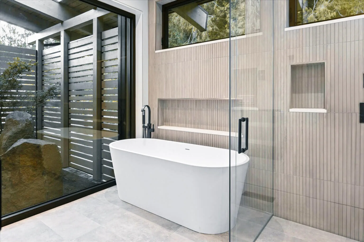 A freestanding bathtub is positioned beside a glass wall looking out onto a fenced outdoor area with natural elements.