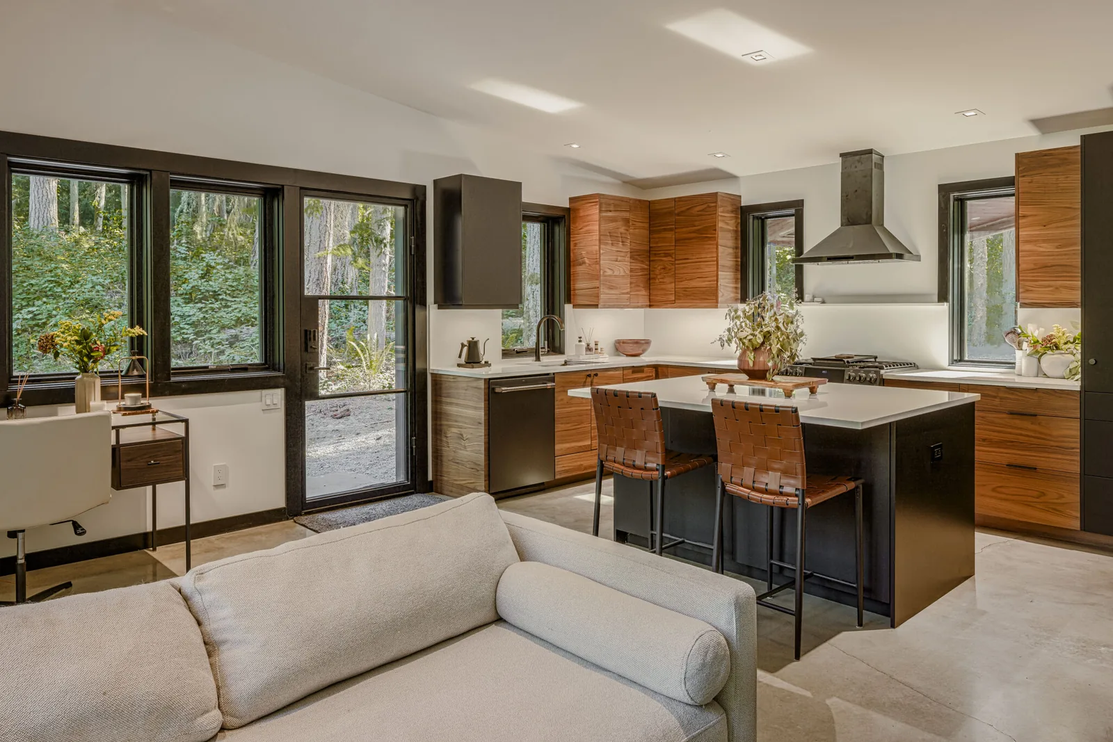 Modern kitchen with wood cabinets, island seating, and black window frames overlooking forested landscape.