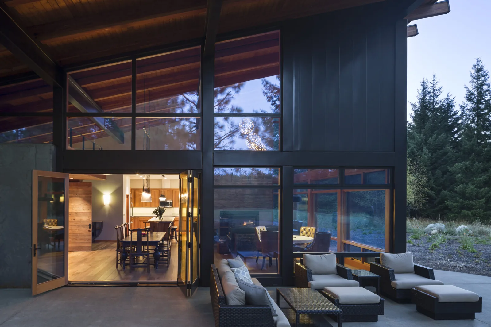 Modern home exterior with large floor-to-ceiling windows and outdoor seating adjacent to an open dining room and kitchen area.