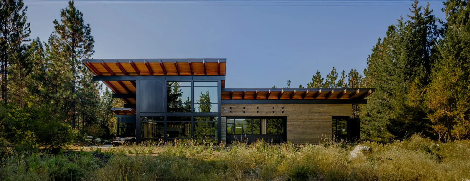 Exterior front view of a modern single-story house with wood and black metal siding, large roof overhangs, and floor-to-ceiling windows.