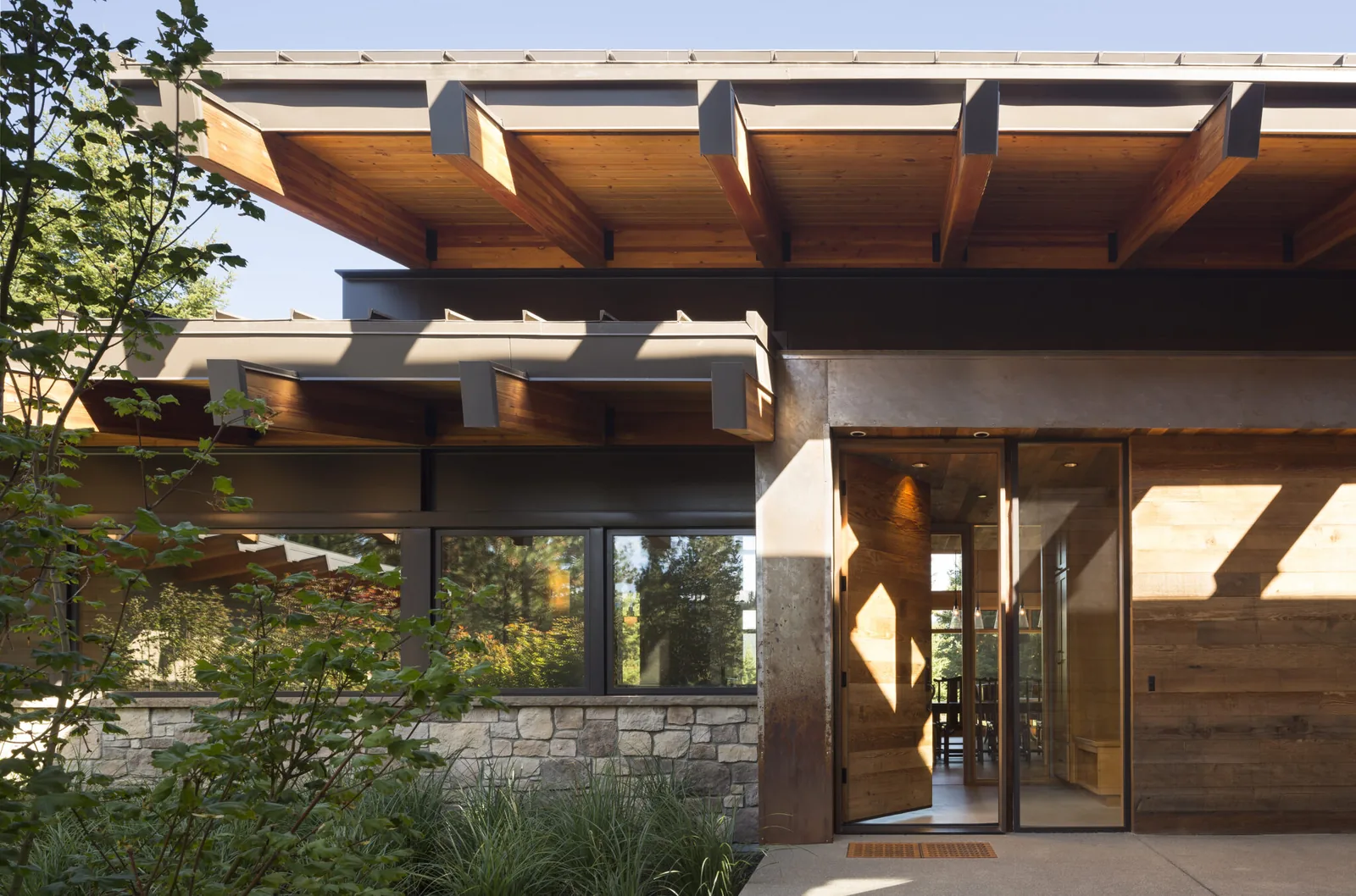 Modern house exterior with exposed wood beams, stone cladding, and a glass entrance door under a covered patio.