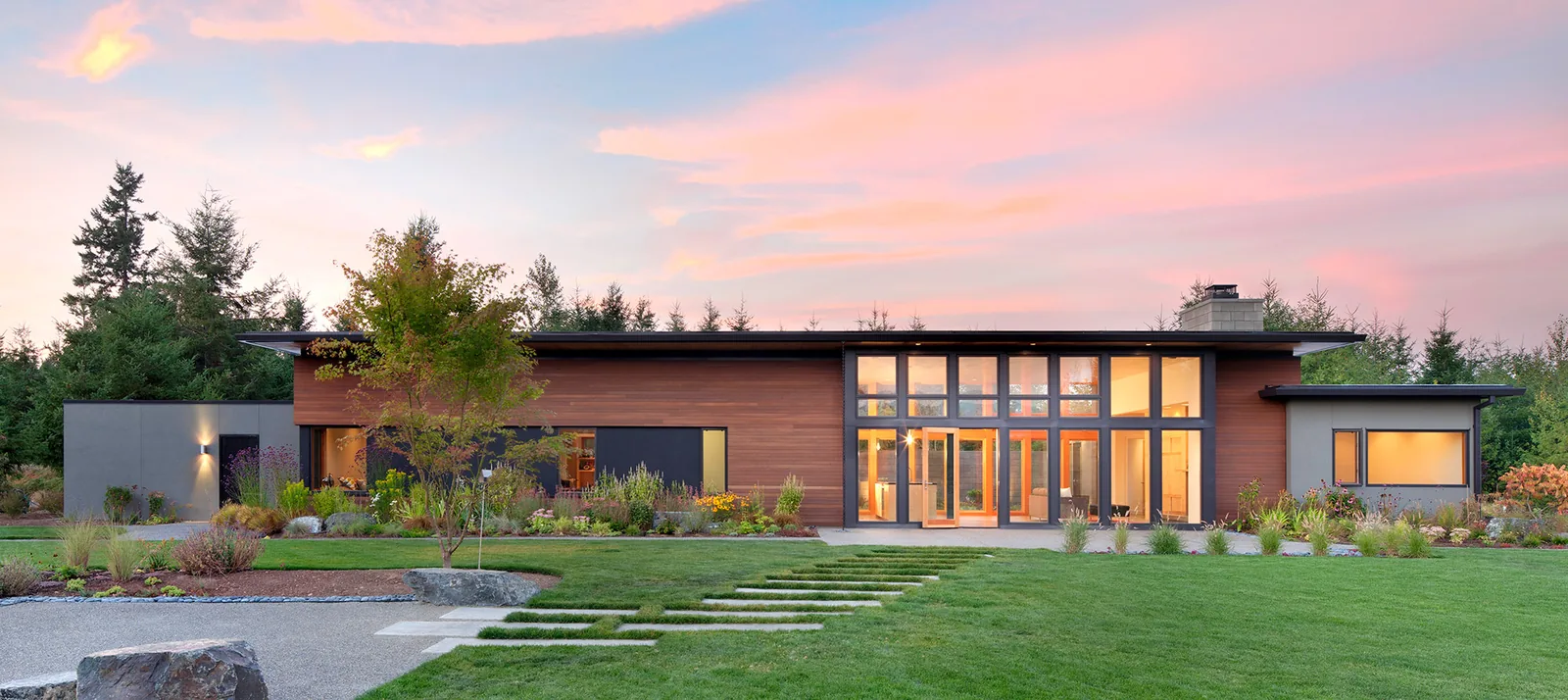 Front exterior view of a modern residential house with wood and stucco facade, large glass windows, and landscaped lawn during sunset.
