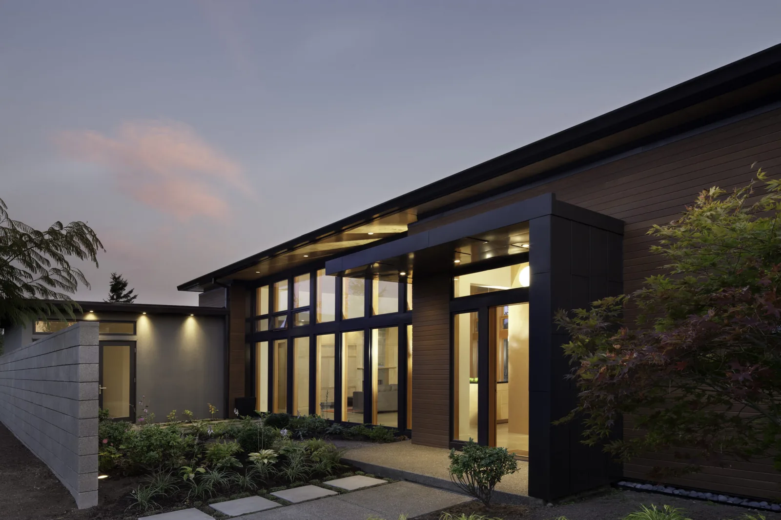 Modern residential house exterior with floor-to-ceiling glazing and a covered entry under a sloped roof at dusk.