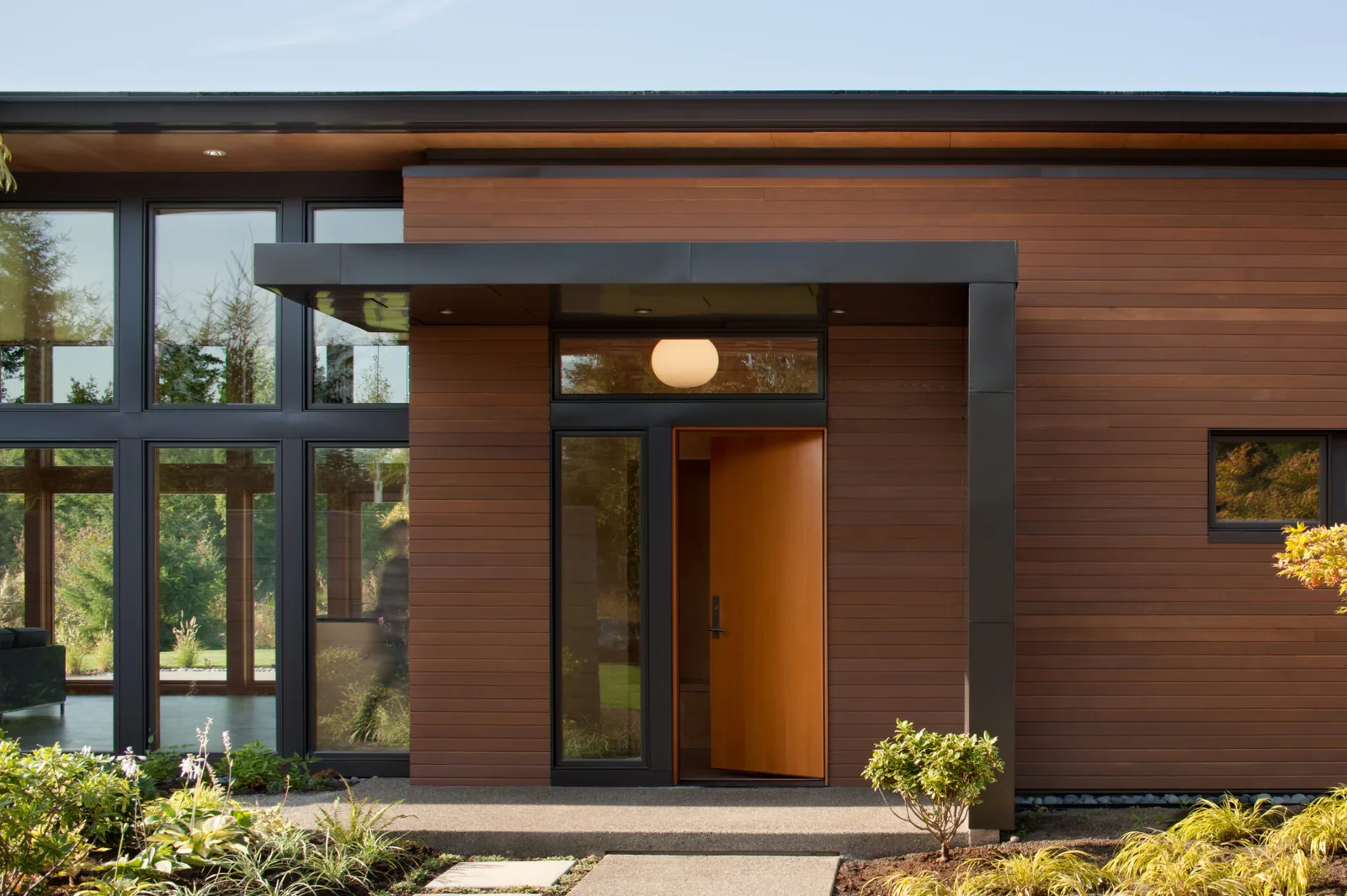 The front entry features wood siding, a pivot door, and expansive black-framed windows under a flat metal canopy.