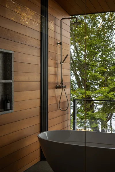 An outdoor shower and freestanding tub with wooden wall cladding and a glass railing overlooking trees.