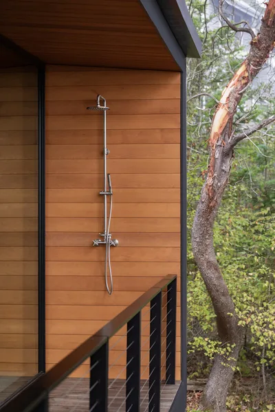 Outdoor shower installed on wood siding wall with metal railing on adjacent balcony, surrounded by natural trees.