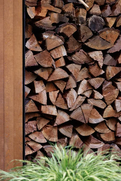 Firewood stacked neatly next to a wood-clad exterior wall with greenery in the foreground.
