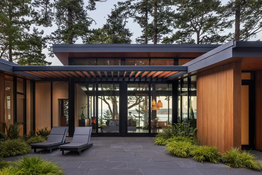 Modern residential exterior featuring floor-to-ceiling glazing, wood siding, and a covered patio area with outdoor seating.