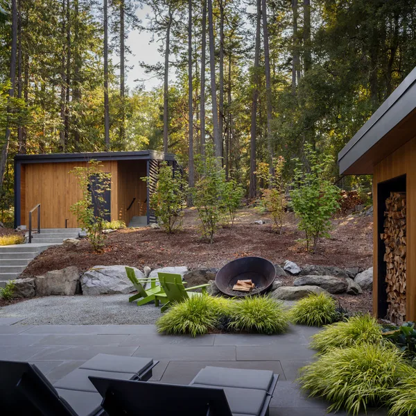 Outdoor living space featuring a fire pit with Adirondack chairs and wood storage sheds on a wooded site.