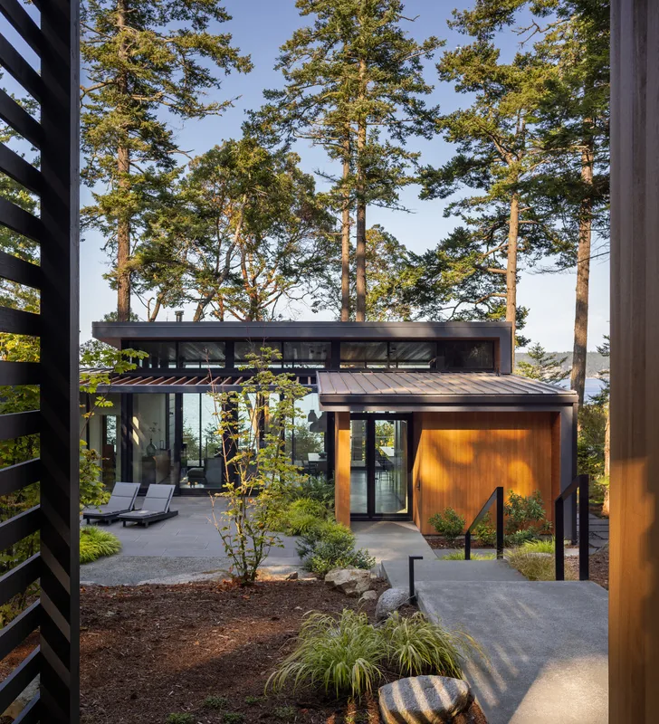 View of a modern single-story house with wood siding and extensive glass walls set in a wooded landscape.