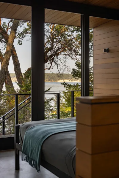 Bedroom with large black-framed windows overlooks natural waterfront landscape.