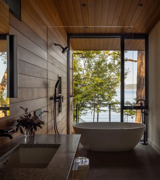 A bathroom with a freestanding tub positioned to face large windows with views of a waterfront and surrounding trees.