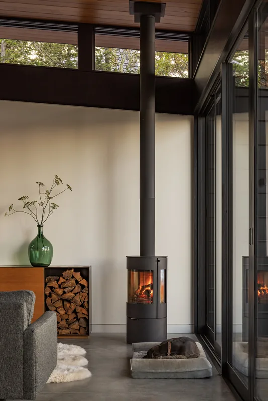 A modern wood-burning fireplace with metal chimney pipe is installed near a glass wall and wood storage inside a living area.