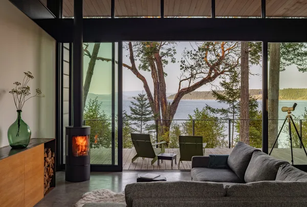 Living room with floor-to-ceiling glazing and a wood stove overlooking a forested waterfront view.