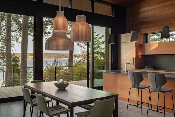 Dining and kitchen space with pendant lighting overlooking a forested water view through floor-to-ceiling windows.