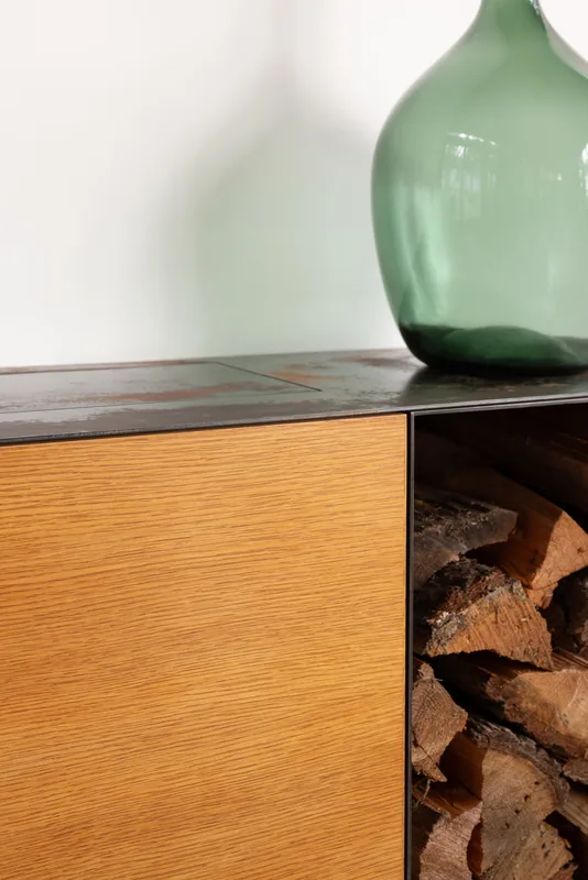 Detail of wood storage with firewood and a green glass vase on surface above.