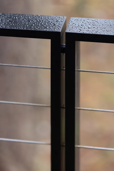 Black metal railing post with cable wire infill and water droplets on top surfaces.