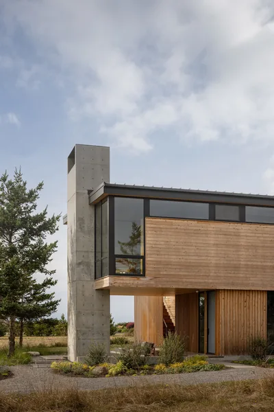 Exterior view of a modern wood and concrete house featuring large windows and a prominent concrete chimney.