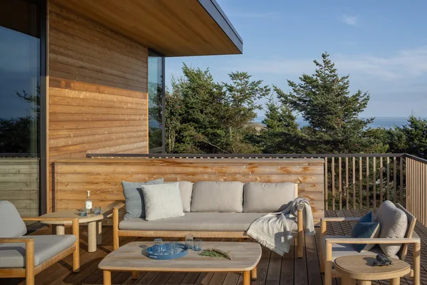 Wood deck patio with cushioned seating and wooden tables overlooking trees and ocean.