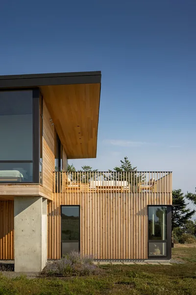 Exterior side view of a two-story contemporary house featuring wood siding, floor-to-ceiling glazing, and an upper-level balcony with vertical wood railings.