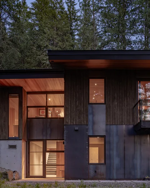 The exterior of the two-story modern house shows wood and concrete materials with large rectangular windows lit from inside at dusk.
