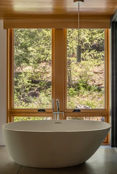 Freestanding bathtub by large wood-framed windows in a bathroom with natural light.