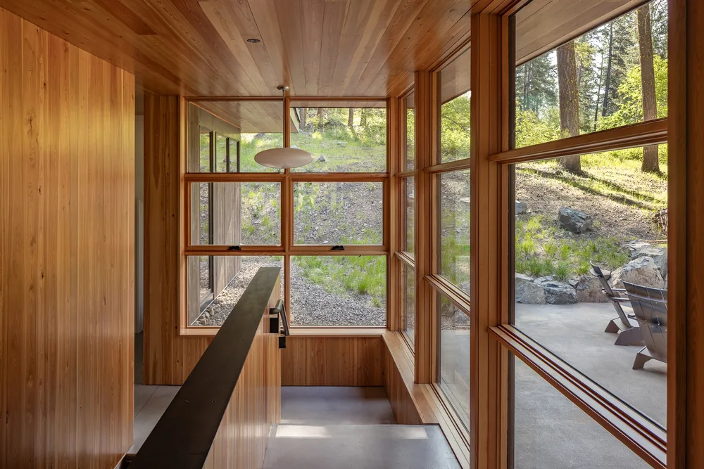 Wood-paneled interior stairwell featuring expansive wood-framed windows overlooking a natural hillside landscape.